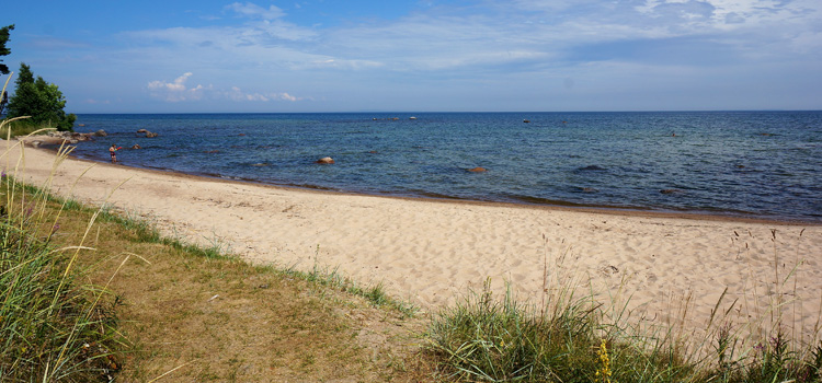 Bilden visar en solig sandstrand vid en sjö. I sjöar som är varma finns det risk att drabbas av badklåda.